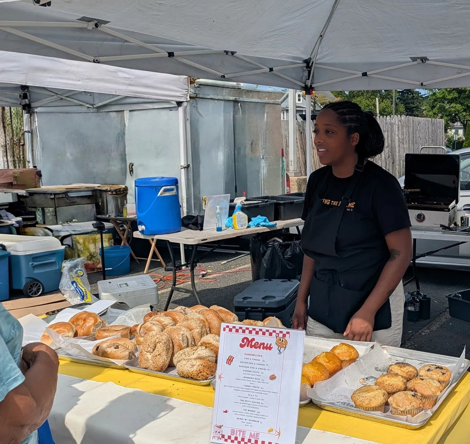 Gabrielle Gilles behind the counter of her Bite Me Bakery stand, trays of bagels and muffins in the foreground