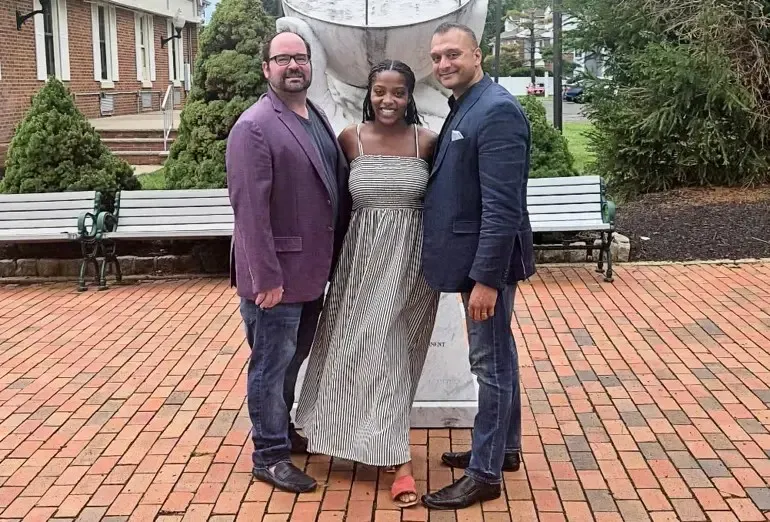 Brian Johdos, Gabrielle Gilles, and Romel D'Munda standing together on a brick plaza in front of a stone monument